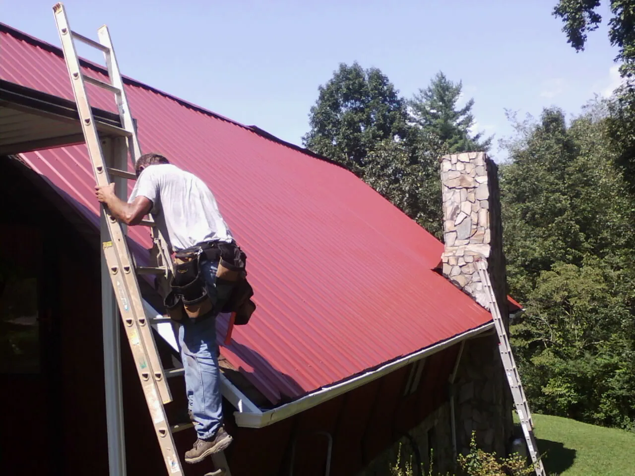 Roofer on a ladder beside a red metal roof and stone chimney — Soffit & Fascia Repair by Roofing Brothers in Seekonk